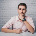 Portrait of a confident young man in a pink shirt sitting at a desk with a brick wall background.